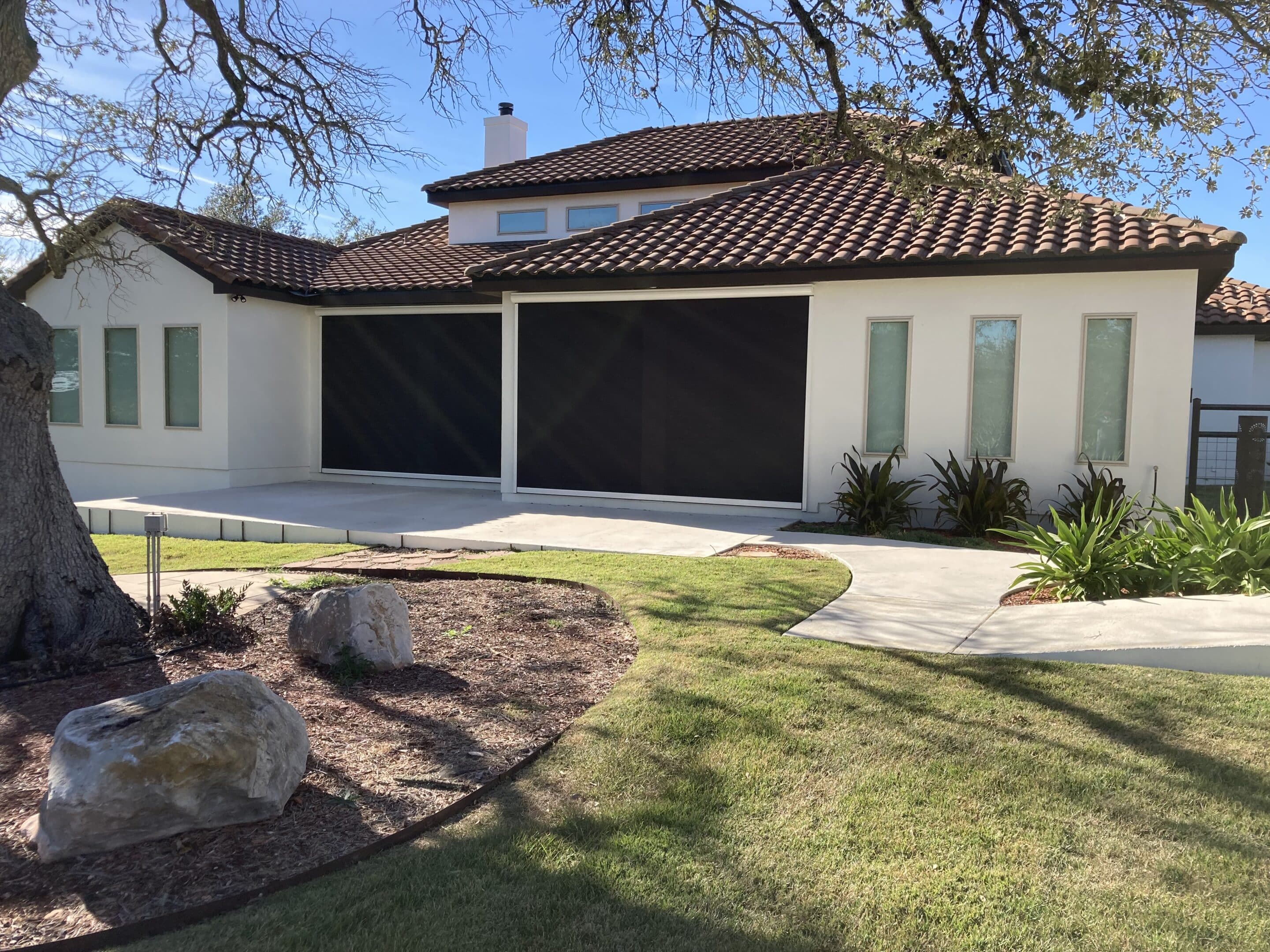 Modern white home with two large black exterior shades covering the patio openings, surrounded by a well kept lawn, pathway, rocks, and landscaped plants under a bright blue sky.