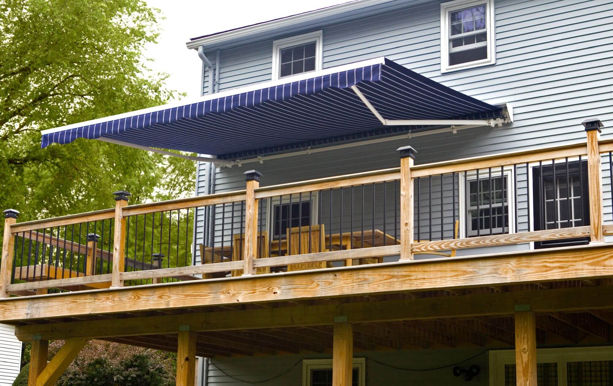 Retractable blue and white striped awning extended over a raised wooden deck on a two story home, shading an outdoor dining table and chairs.