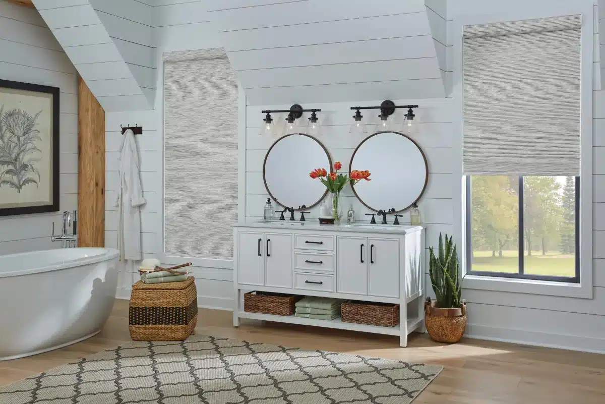 Light textured roller shades in a bright bathroom with a freestanding tub, white double vanity, round mirrors, woven baskets, and wood flooring.