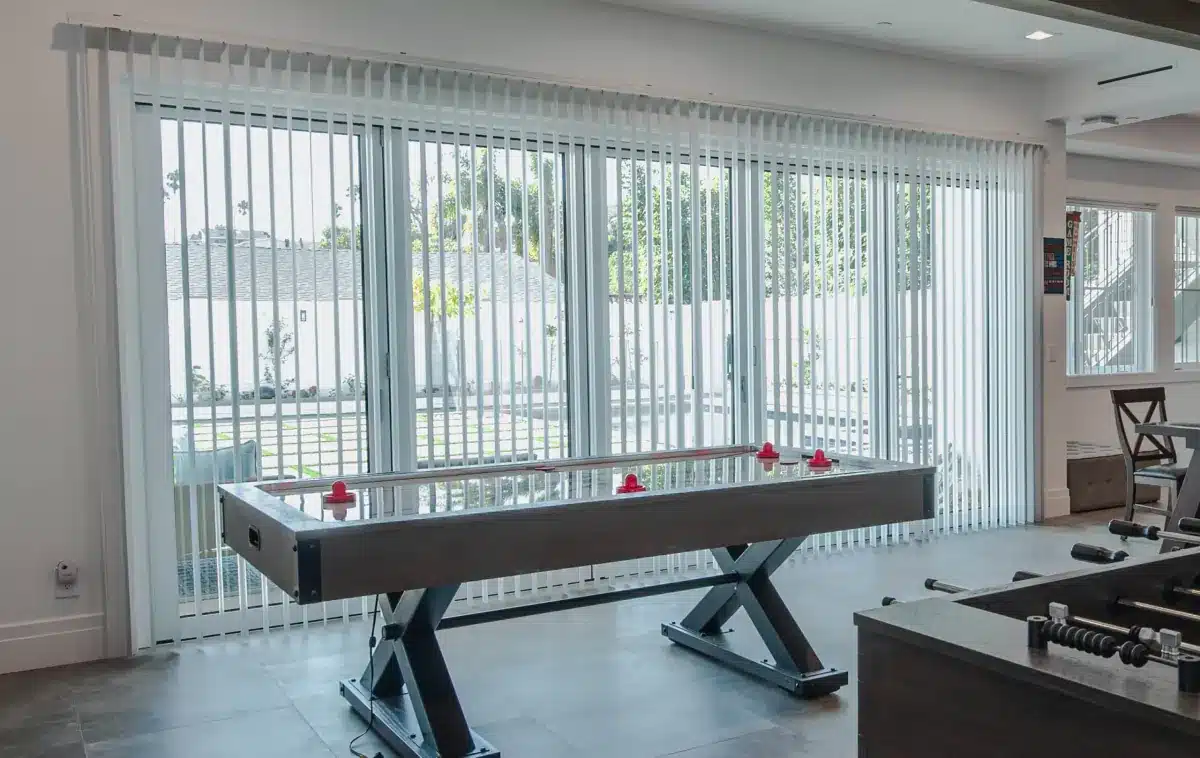 White vertical blinds frame floor-to-ceiling glass doors overlooking a patio in a game room in Venice, FL