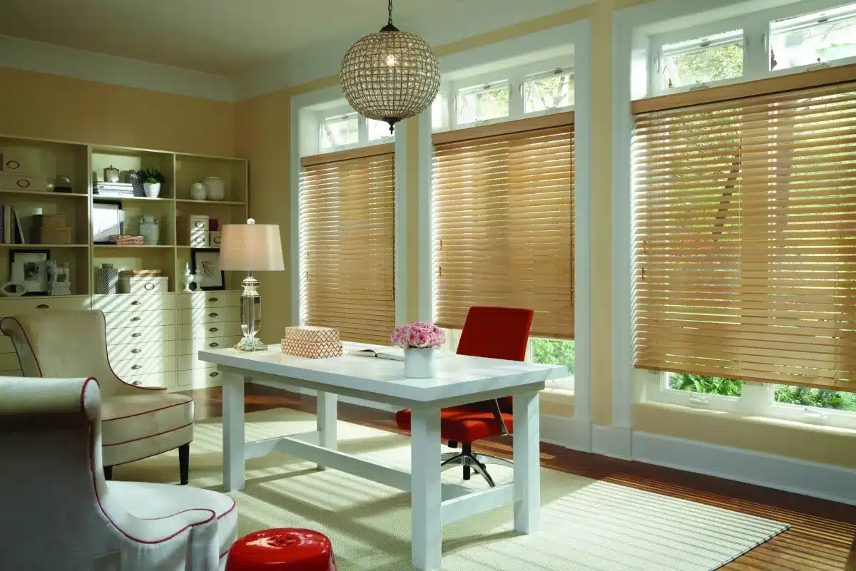 Wood blinds in natural tone frame large windows in a bright home office with white desk and red chair in Venice, FL
