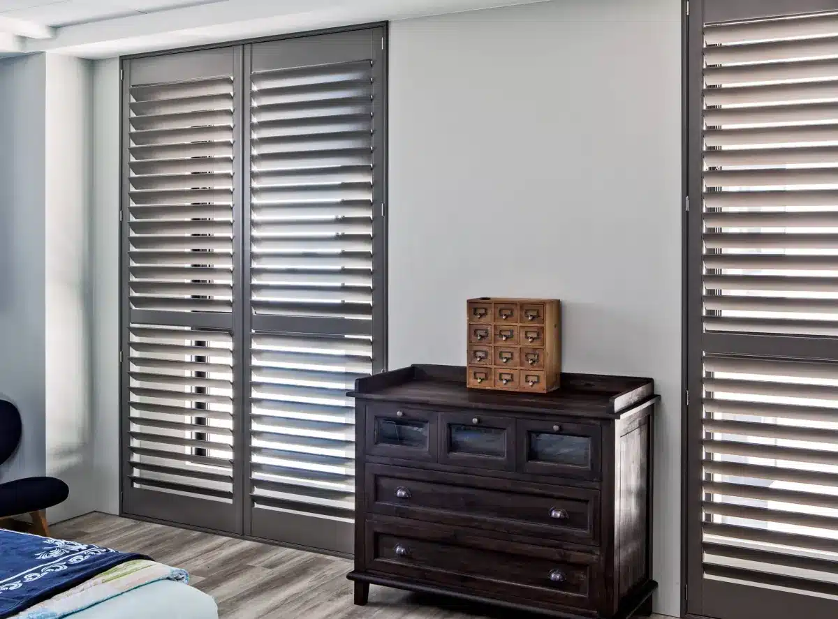 Dark plantation shutters frame a bedroom window above a wooden dresser with decorative storage box in Venice, FL