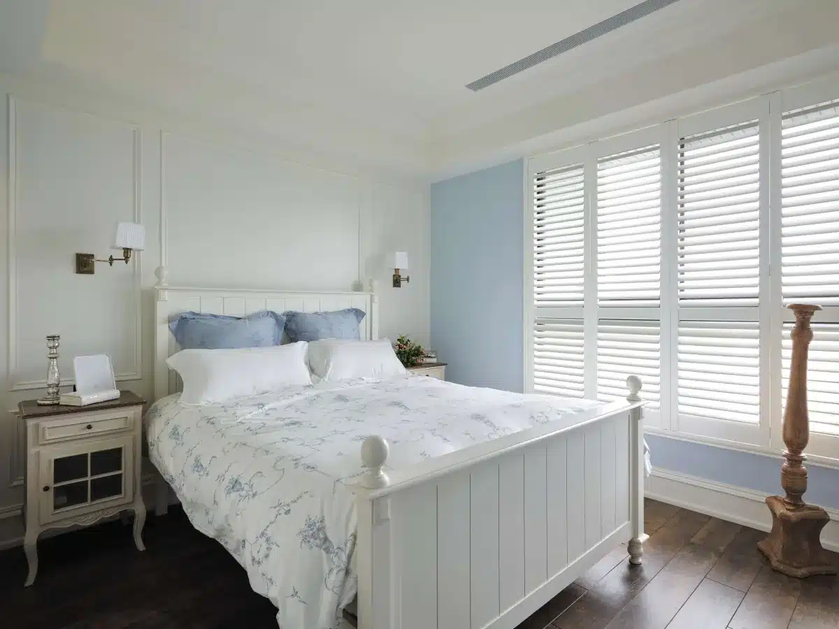 White plantation shutters frame large bedroom windows with blue accent wall and traditional furnishings in Venice, FL