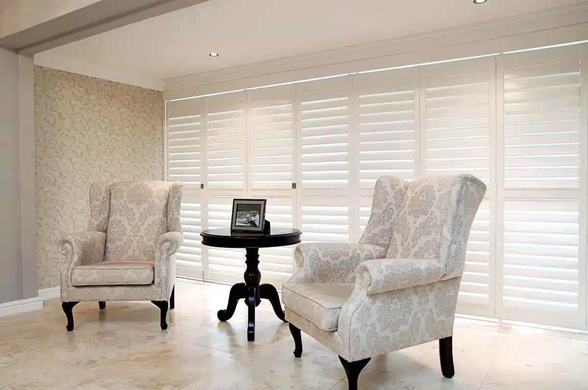 White plantation shutters frame a living room seating area with two patterned wingback chairs and a side table in Venice, FL